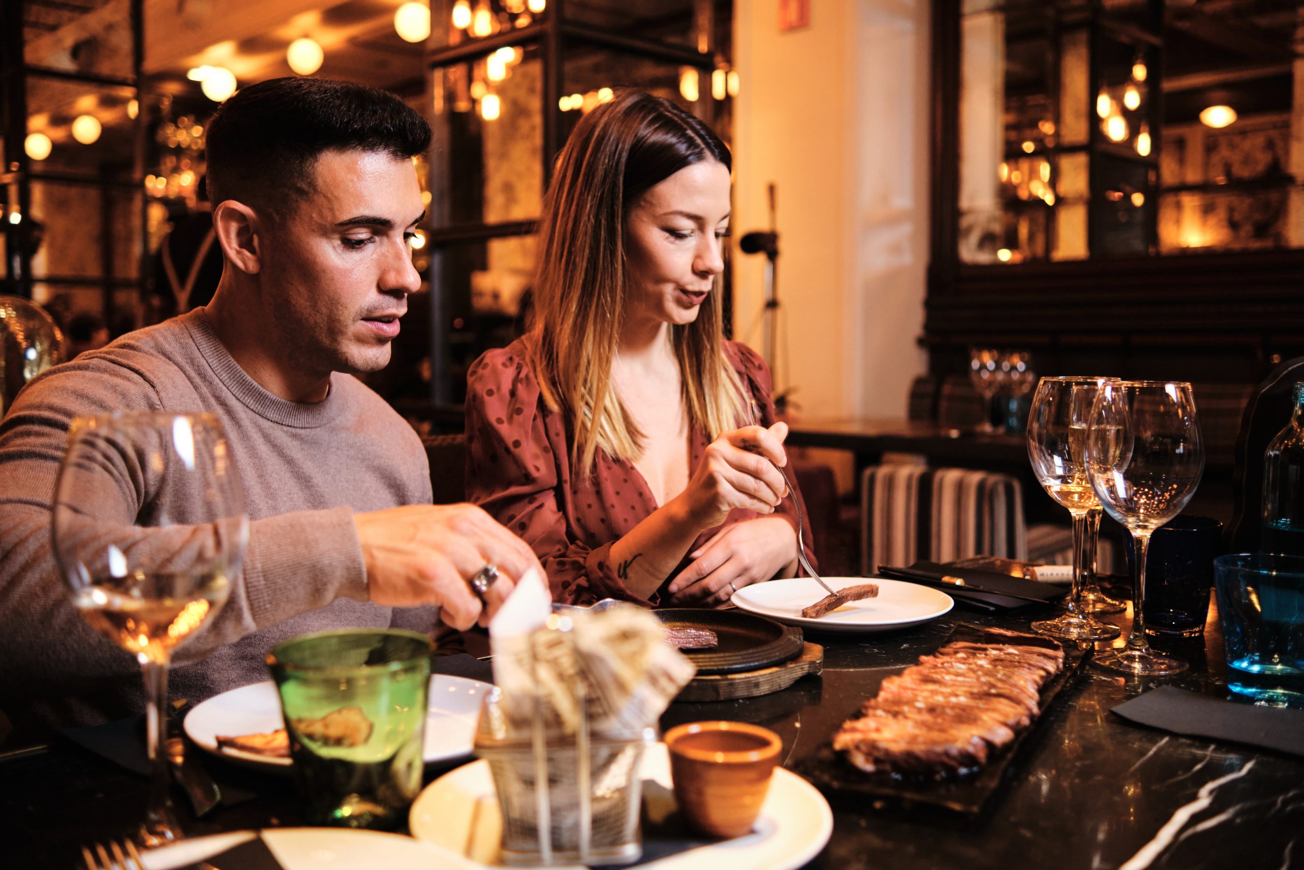 Portrait of young couple enjoying time together and having dinner on a date at fancy restaurant. Couple eating with glasses of wine on table.